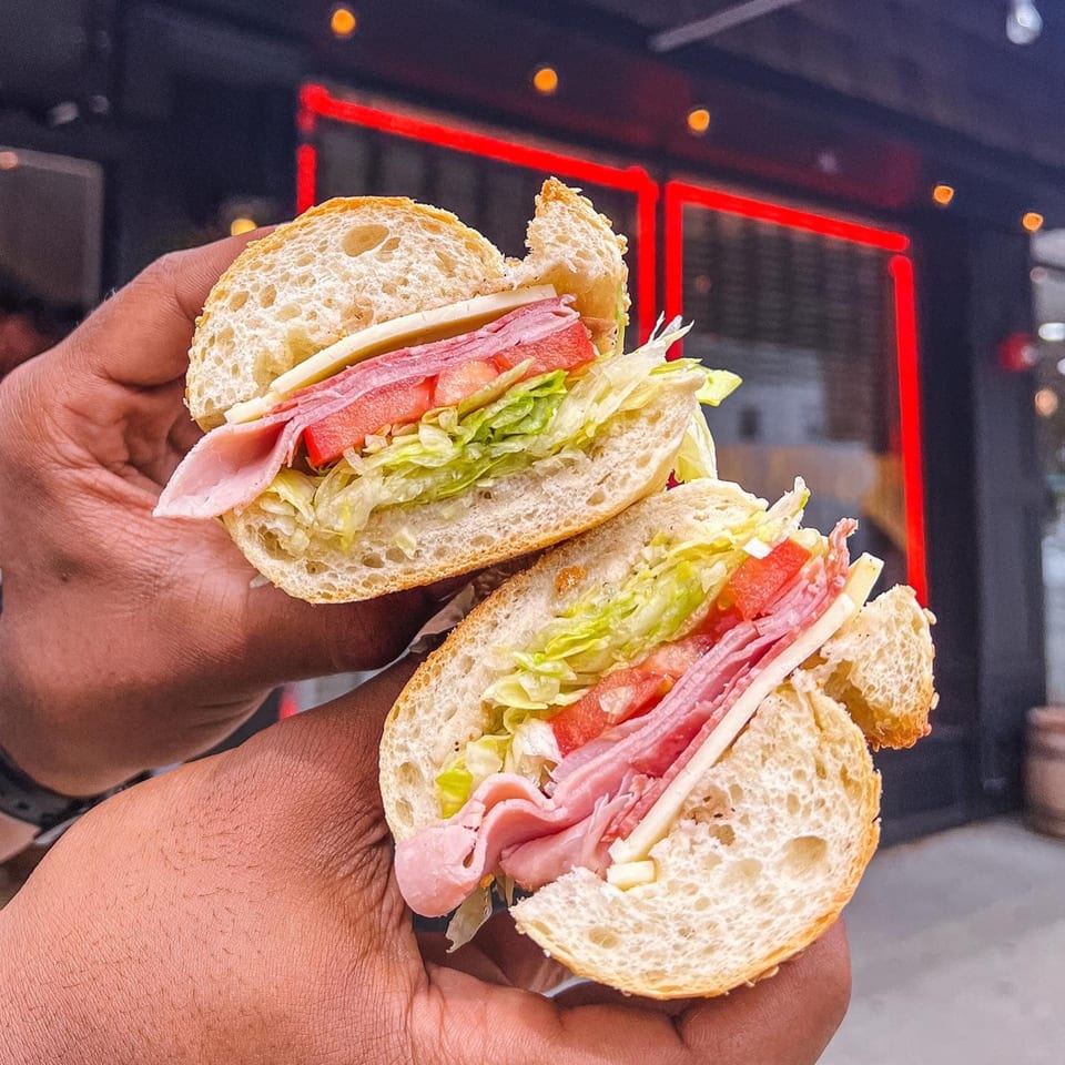 Hands holding a sliced sub sandwich filled with ham, salami, cheese, lettuce, and tomatoes.