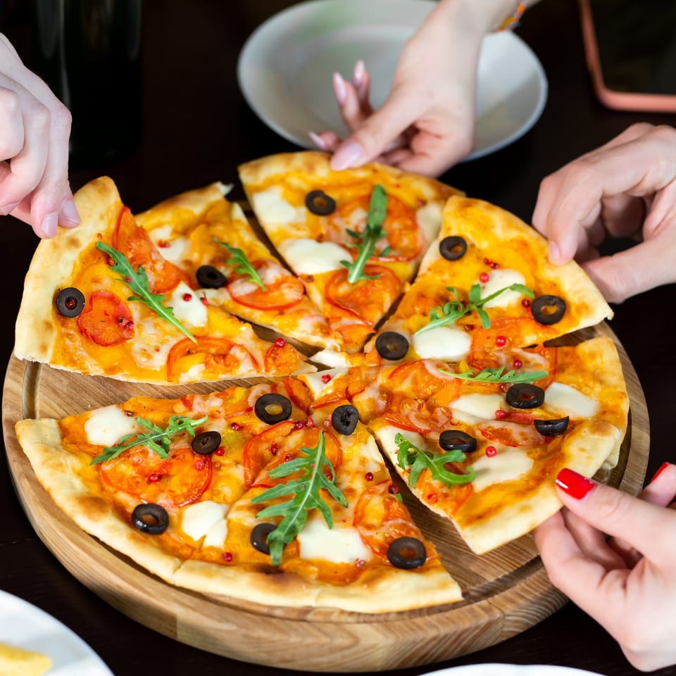 Hands reaching for slices of Soho Pizza & Grill's pizza with tomatoes, olives, mozzarella, and arugula.