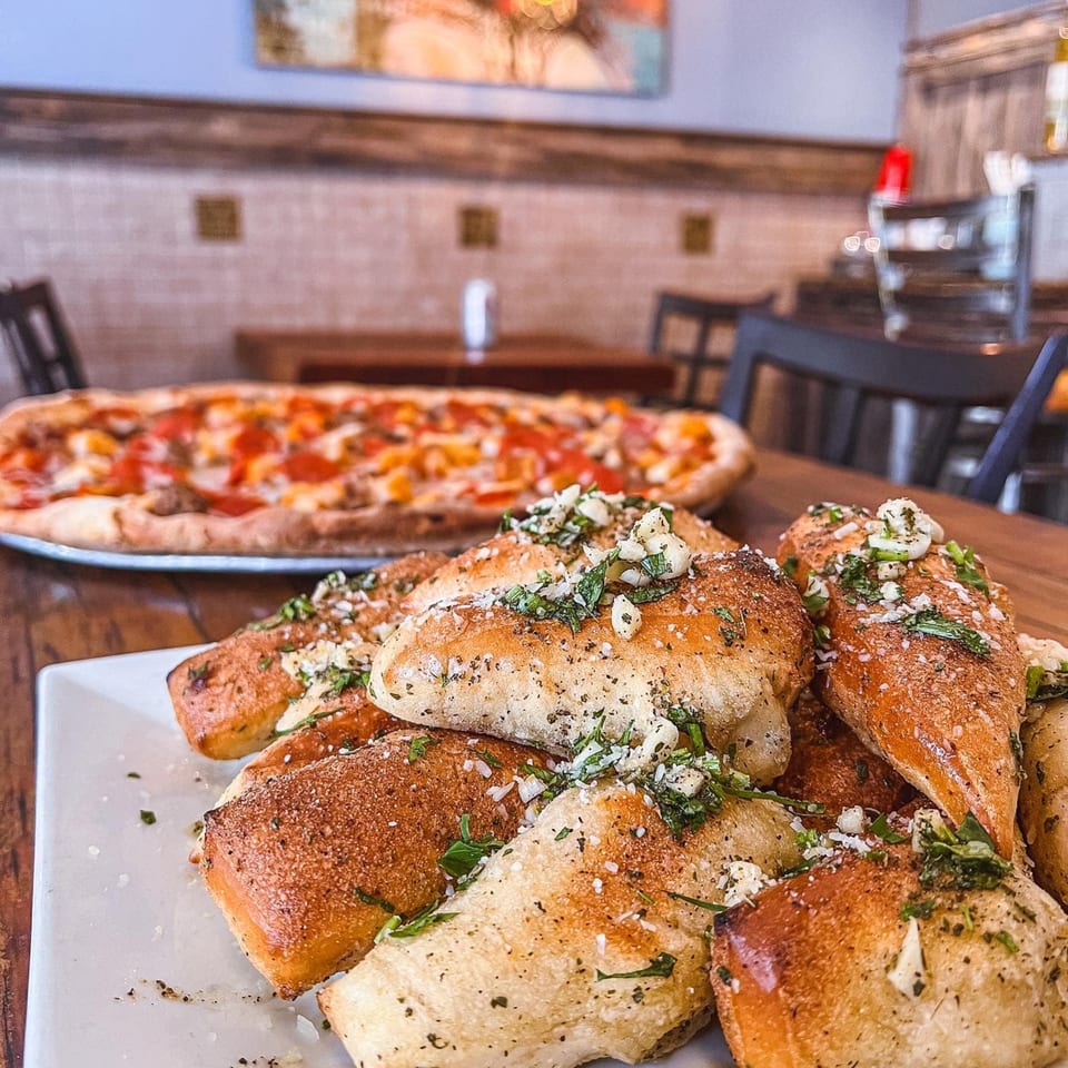 Garlic knots with herbs and parmesan on a white plate, with a pizza in the background.